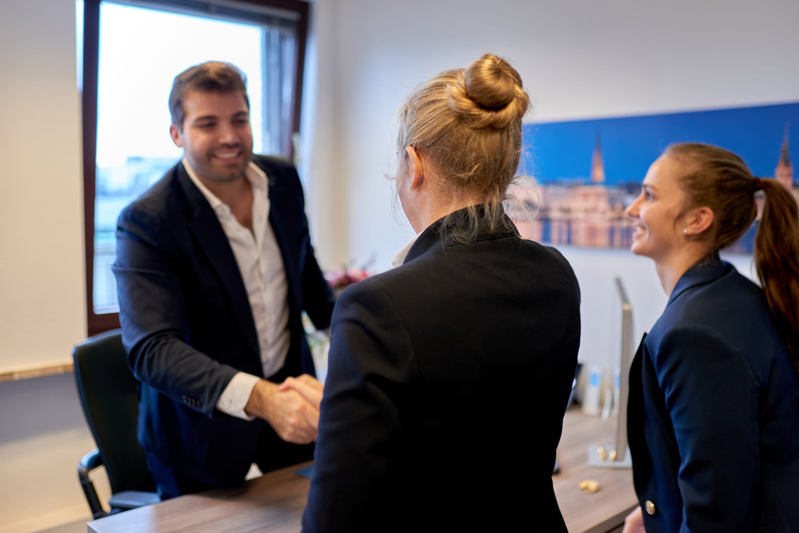 Immobilienmakler begrüßt Kundin mit Handschlag am Schreibtisch, im Hintergrund ein Hamburg-Panoramabild an der Wand